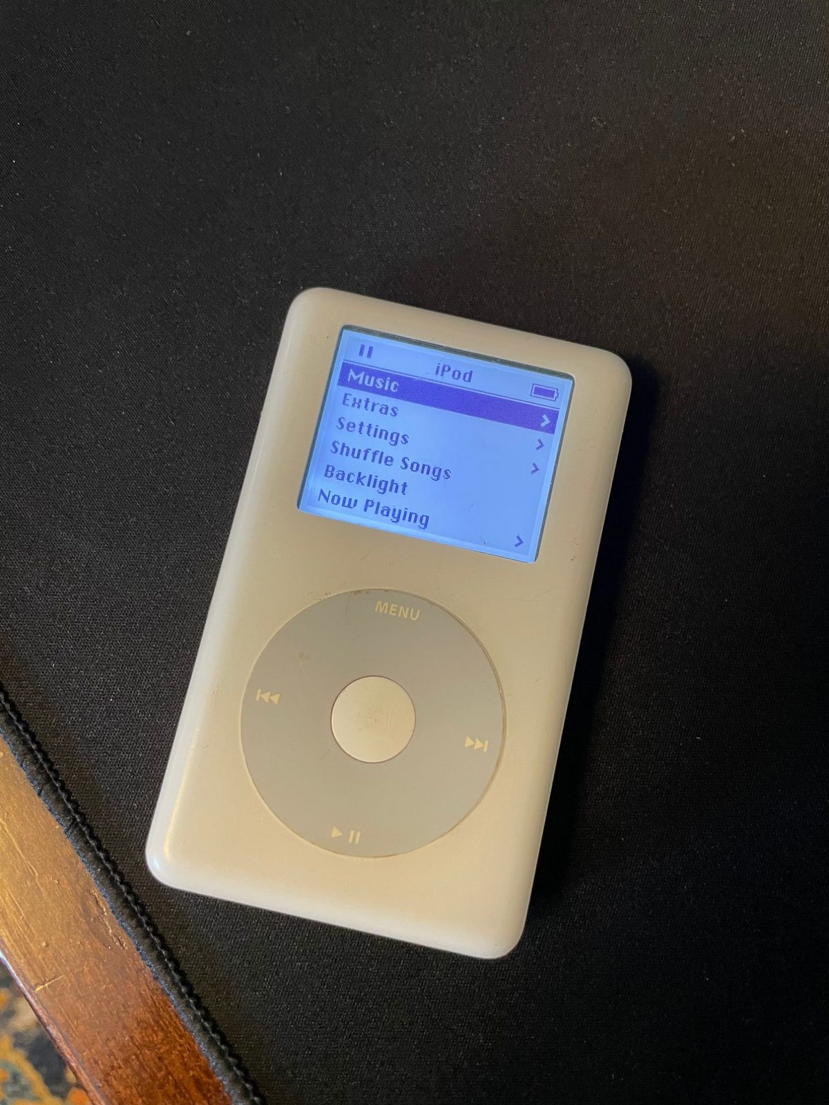 White fourth-generation iPod with a grayscale click wheel lying on dark fabric, powered on. The monochrome screen shows the main menu — Music, Extras, Settings, Shuffle Songs, Backlight, Now Playing — with a pause icon and a full battery indicator in the status bar.