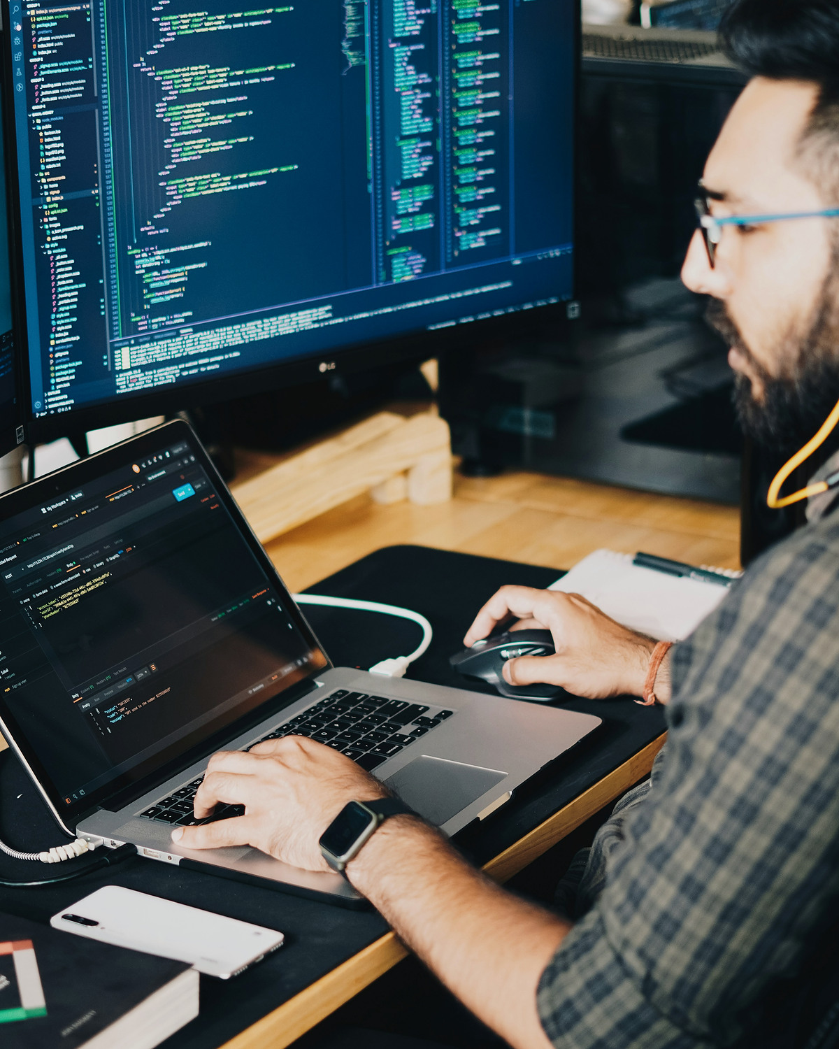 A software developer working across a code-filled monitor and laptop.